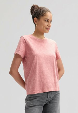 Junge Frau mit lockigem Haar, zu einem Dutt gebunden, trägt ein rosa kurzärmeliges Shirt und graue Jeans, steht mit den Händen hinter dem Rücken und schaut nach rechts.