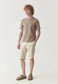 Tricou din bumbac de culoare bej deschis, pantaloni scurți din in crem și sandale verde măsliniu. Modelul stă pe un fond simplu, evidențiind potrivirea ținutei.
