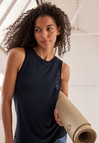 Top senza maniche di colore blu navy scuro con scollo a U e un delicato motivo testurizzato, abbinato a un tappetino da yoga beige arrotolato tenuto sotto il braccio.