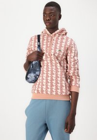 Felpa con cappuccio a motivo geometrico rosso su sfondo rosa chiaro, abbinata a pantaloni della tuta blu e una piccola borsa a tracolla blu tie-dye.