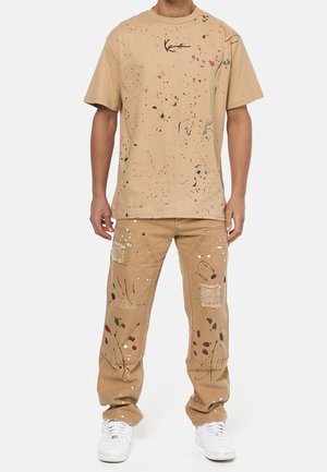 Camiseta de algodón beige con un diseño de salpicaduras de pintura multicolor y un logotipo; combinada con pantalones beige que presentan parches y un detalle de salpicaduras similar.