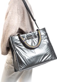 Grand sac à main métallique gris avec des anses tressées multicolores et une texture brillante, présentant une forme rectangulaire et des détails de couture visibles.