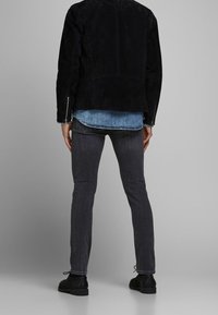 Černá semišová bunda přes svetle modrou denimovou košili, šedé slim-fit džíny a černé boty. Viditelné švy a zipy na bundě.