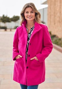 Cappotto di lana fucsia con colletto, bottoni frontali e tasche laterali. Indossato sopra una camicetta a fantasia bianca e nera, abbinato a jeans blu.