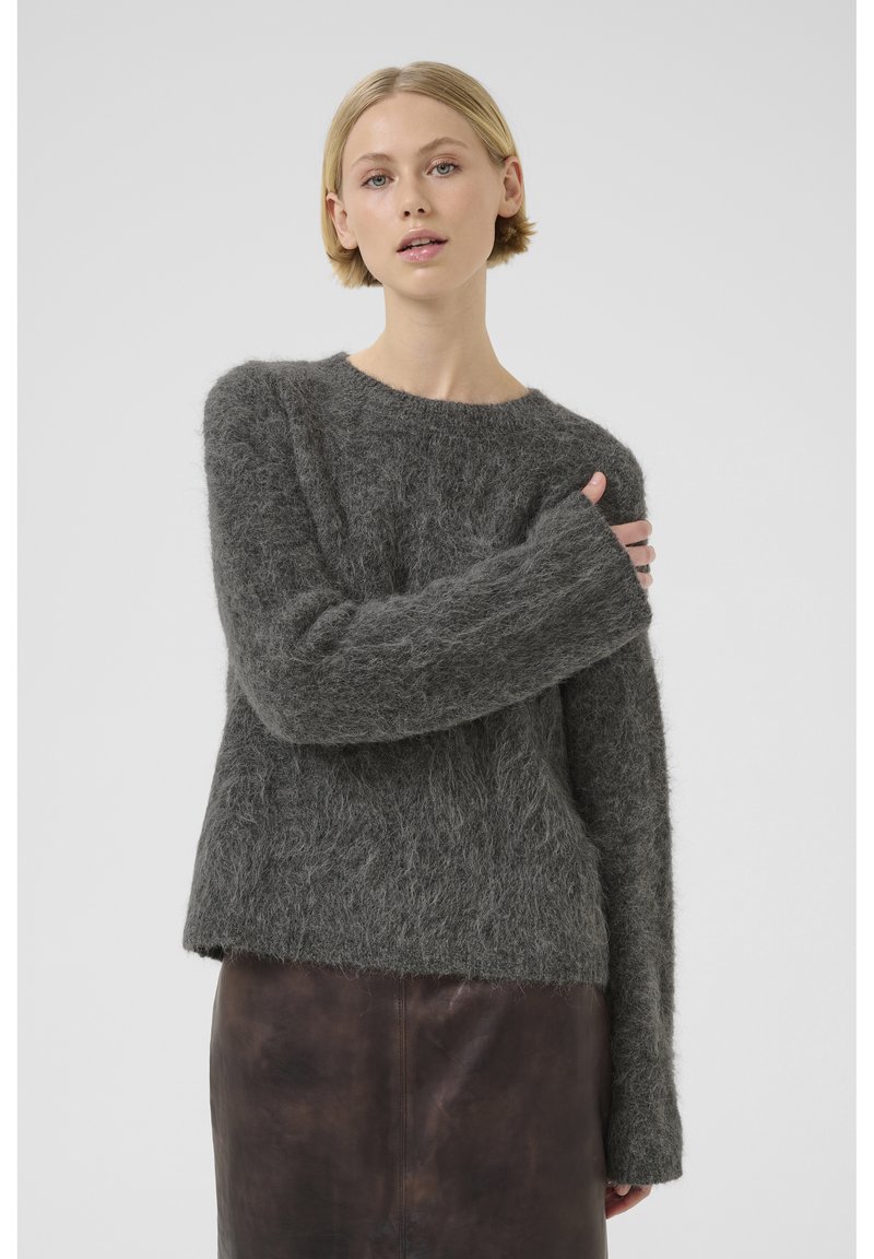 Pull en laine gris flou en matériau texturé, avec un col rond, des manches longues et un ourlet légèrement court. Associé à une jupe en cuir noire.