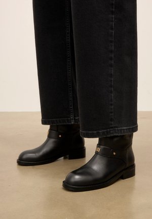 Bottines en cuir noir à talons bas portées avec un jean noir à jambes larges sur un sol marron clair.