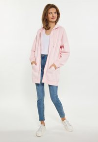 Sudadera oversized rosa claro con cremallera, cordones ajustables y dos bolsillos frontales; combinada con jeans skinny azul claro y zapatillas blancas.