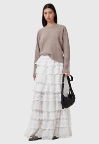 Beige ribstrikket sweater med en afslappet pasform, parret med en hvid lagdelt blonde-nederdel med bølgede kanter. Sort skuldertaske og mønstrede sko.