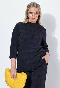 Top a quadri navy con colletto alto, maniche a tre quarti e tessuto texturizzato; abbinato a una borsa di paglia gialla per un contrasto.