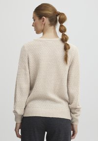 Maglione lavorato a maglia beige chiaro con un motivo testurizzato, orlo a costine e maniche lunghe; visto da dietro, abbinato a pantaloni scuri.