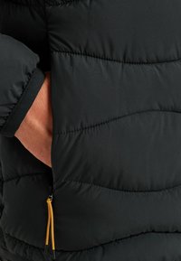 Crna puf jakna s kvadratnim uzorkom, s elastičnom manžetom i vidljivim žutim povlačnim trakama na bočnoj džepu za podešavanje.