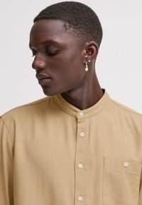Beige skjorte med knapper og stiv krage, med brystlomme og glatt tekstur. Modellen bærer en perleøredobb.