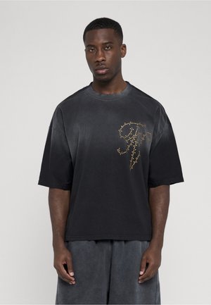 Jonge man met een loszittend zwart T-shirt met goudkleurige studs en letters op de borst en bijpassende donkere korte broek, staand tegen een effen achtergrond.