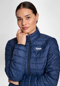 Granatowa kurtka puchowa z zapięciem na zamek i wysokim kołnierzem, z przeszywanym pikowanym wzorem oraz logo na piersi. Gładkie wykończenie materiału.