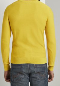 Żółty sweter z prążkowanej dzianiny z długimi rękawami, okrągłym dekoltem i dopasowanym wykończeniem, w połączeniu z niebieskimi dżinsami z ozdobnym szwem na kieszeniach.