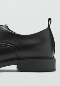 Zapato negro de cuero con textura suave, forma redondeada, tacón bajo y una lengüeta en la parte trasera. Presenta detalles de costura sutiles.