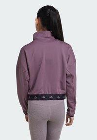 Chaqueta pullover morada recortada con cuello alto y etiqueta AEROREADY. Presenta una cintura elástica negra con acentos de logo y mangas con puños.