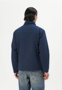 Navy blue jacket with a collar, visible stitching details, and a relaxed fit. Worn with light blue denim jeans featuring back pockets.