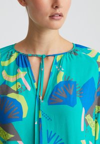 Blouse colorée avec un fond turquoise et un mélange de motifs abstraits en vert, bleu, gris et rose. Détail d'encolure en clé.
