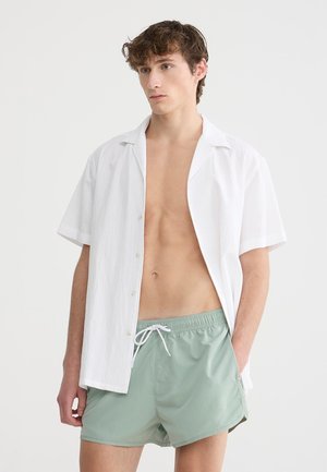 Camicetta bianca a maniche corte con tessuto testurizzato, aperta al colletto, abbinata a pantaloni da bagno verde menta con vita elasticizzata e stringa.