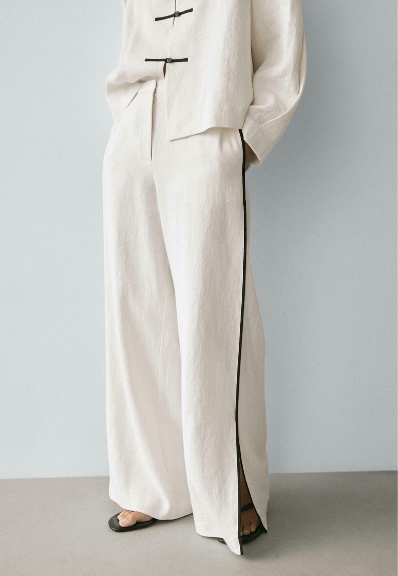 Pantalon ample couleur blanc cassé avec bande noire sur les côtés et fente devant, porté avec un haut assorti et des sandales noires à bout ouvert.