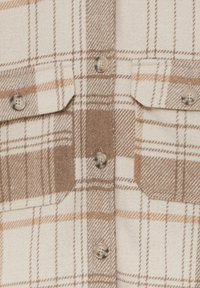 Gros plan sur un tissu à carreaux beige et marron avec deux poches poitrine et des boutons beiges au centre.