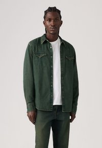 Πράσινο τζιν πουκάμισο με μυτερό γιακά, διπλές τσέπες στο στήθος και κούμπωμα με κουμπιά, φορεμένο πάνω από λευκό t-shirt, σετ με τζιν παντελόνι στο ίδιο πράσινο χρώμα.