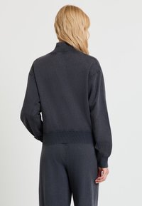 Blonde Person von hinten, trägt ein dunkelgraues Sweatshirt mit langen Ärmeln und passende Hose vor einem einfarbigen hellen Hintergrund.