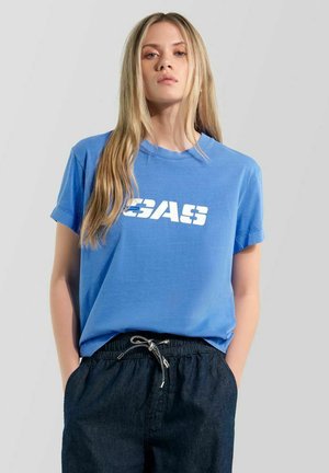 Giovane donna con lunghi capelli biondi che indossa una maglietta blu con testo bianco e pantaloni scuri con coulisse, in piedi davanti a uno sfondo semplice.