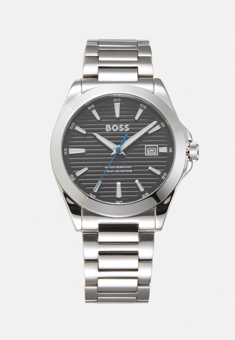 Sølv rustfritt stål armbåndsur med en svart teksturert urskive, lysende timeindekser, blå sekundviser og datovisning; vanntett opptil 50 meter.