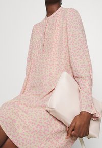 Sukienka w panterkę w jasnoróżowym i fioletowym kolorze, z wysokim dekoltem, detalami z guzikami i długimi rękawami; trzyma gładką, jasnoróżową kopertówkę.