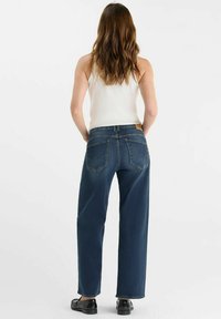 Donkerblauwe wijde jeans met achterzakken die een opvallend naaddesign hebben, gecombineerd met een getailleerde witte tanktop en zwarte schoenen.