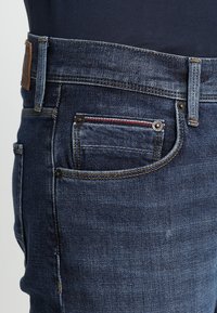 Primer plano de un bolsillo de unos pantalones de mezclilla azul oscuro, con un lazo de cinturón visible, costuras y pequeños remaches de metal.