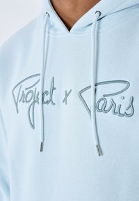 Felpa azzurra con cappuccio e testo ricamato grigio "Project x Paris." Presenta cordini con punte metalliche silver e una texture in tessuto morbido.
