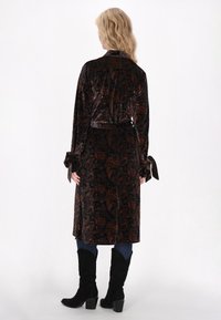 Cappotto lungo in velluto dalle ricche tonalità scure con motivo paisley, vita legata e maniche svasate. Abbinato a jeans blu e stivali neri.