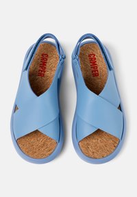 Siniset sandaalit, joissa on ristikkäiset hihnat sileästä materiaalista ja korkkipohja. Takahihna varmistaa mukavan istuvuuden, sisäpuolella punainen "Camper" -logo.