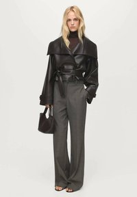 Giacca cropped in pelle marrone con colletto ampio, abbinata a pantaloni grigi a vita alta. La modella tiene una piccola borsa marrone, con scarpe open-toe.