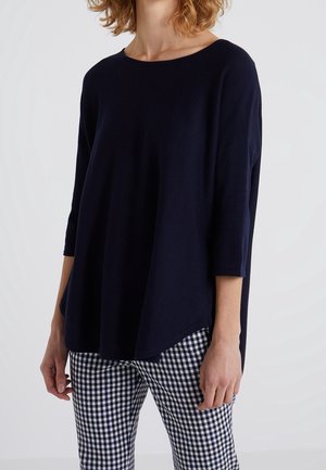 Persoon draagt een losvallende donkerblauwe top met driekwart mouwen en zwart-wit geruite broek, staand tegen een effen achtergrond.