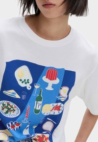 Witte t-shirt met een blauwe graphic met verschillende voedselillustraties, waaronder een taart, drankjes en gerechten. Korte mouwen en een ronde hals.