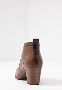 Brązowe skórzane botki do kostki z piętą w klocku i czarną pętelką na białej powierzchni i tle.