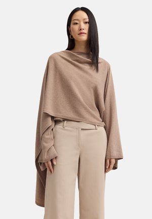 Woman with straight black hair wearing gold hoop earrings, a loose brown poncho-style sweater, and beige pants against a white background.