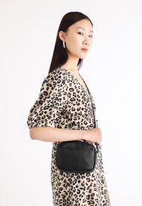 Μαύρη ορθογώνια τσάντα ώμου με υφή, λεπτό λουρί, λείες άκρες, κρατιέται από ένα άτομο που φοράει φόρεμα με λεοπάρ σχέδιο.