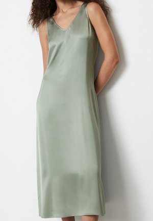 Femme portant une robe nuisette en satin vert clair sans manches, debout contre un mur blanc avec les bras derrière le dos.