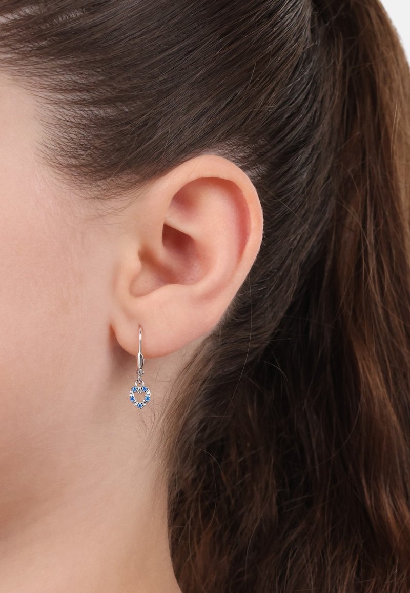 Orecchini a cerchio argento con pendente a forma di cuore, impreziositi da pietre blu incastonate in un design scintillante, indossati all'orecchio di un modello.
