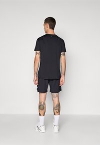 Mann med tatoveringer iført en svart t-skjorte, svarte shorts, hvite sokker og hvite joggesko står vendt mot en enkel lys bakgrunn.