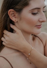 Náušnice zo žltého zlata s kvetinovým dizajnom a zlatý náramok s drobnými perlami, nosené na opálenej ruke na neutrálnom pozadí.