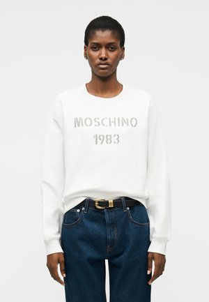 Žena v bílé mikině Moschino 1983, tmavě modrých džínech s vysokým pasem a černém pásku se zlatou sponou na jednobarevném pozadí.