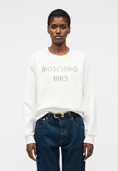 Γυναίκα με λευκό φούτερ Moschino 1983, σκουρό μπλε τζιν ψηλόμεσα και μαύρη ζώνη με χρυσή αγκράφα, ενάντια σε απλό φόντο.