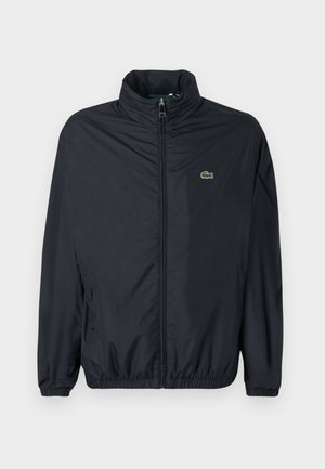 Veste coupe-vent noire avec un col montant, une fermeture éclair à l'avant et des poignets élastiques. Présente un petit logo vert sur la poitrine gauche. Matière légère.