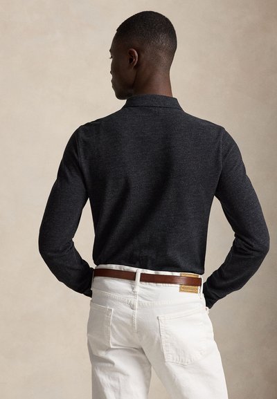 Vyras atsuktas nugara, dėvi tamsią ilgomis rankovėmis palaidinę įkištą į baltas kelnes su rudu odiniu diržu priešais smėlio spalvos foną.